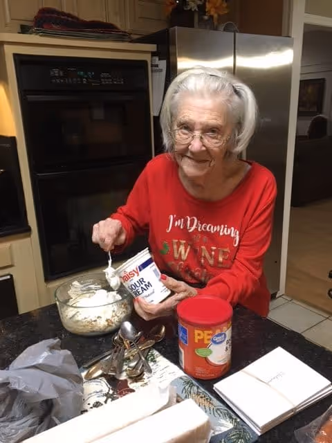 An elderly woman with white hair and glasses wearing a red shirt that says 'I'm Dreaming of Wine' is sitting at a kitchen counter. She is holding a container of sour cream and spooning some into a glass bowl filled with food. Various kitchen items, including a container of peanut butter, spoons, and napkins, are on the counter. The background shows a refrigerator and an oven.