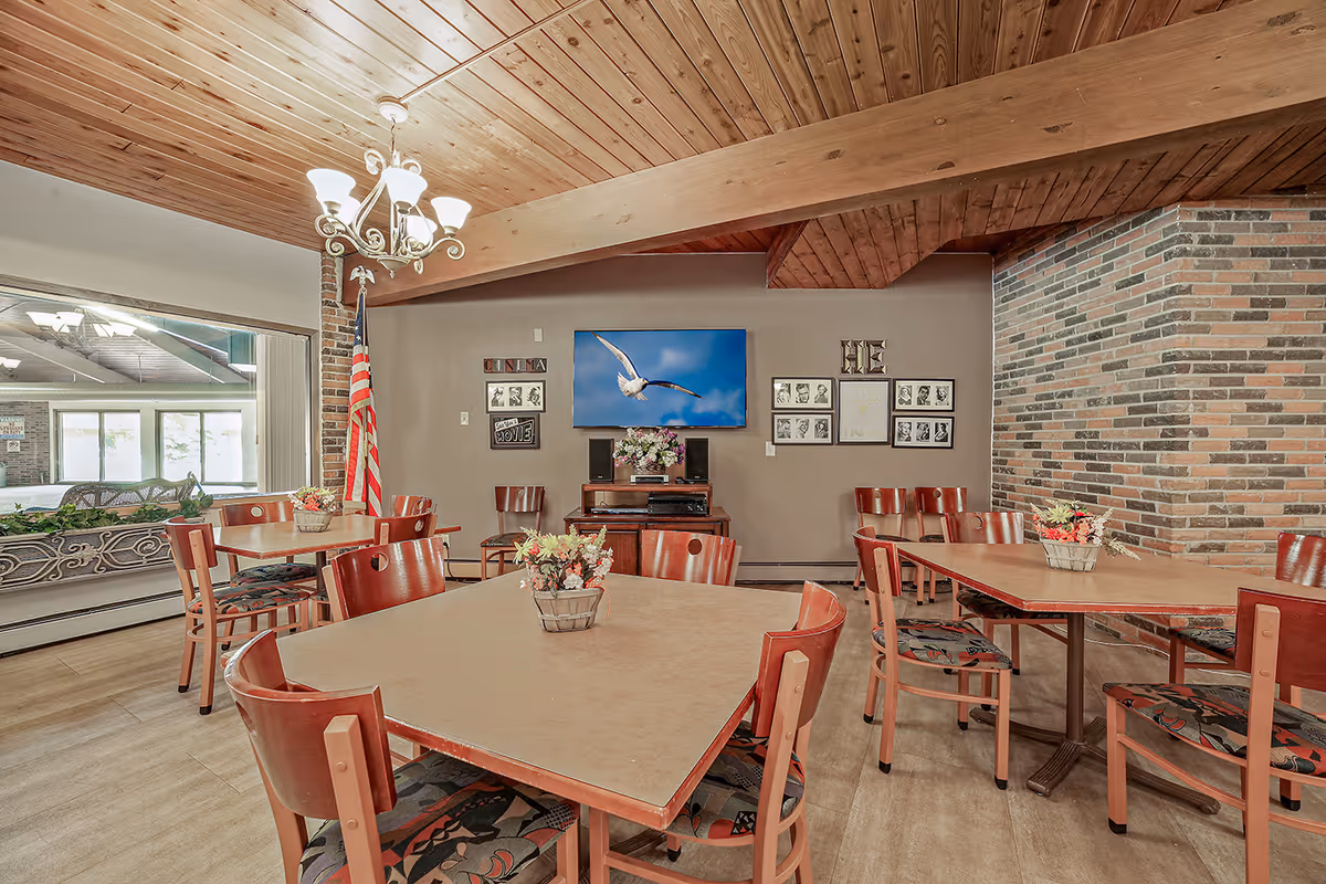 A dining room with multiple wooden tables and chairs arranged neatly. The room features a wooden ceiling with a chandelier, a brick wall with framed pictures, and a flat-screen TV displaying a bird in flight. There is an American flag in the corner and flower arrangements on the tables. A large window shows an adjacent room with more seating.