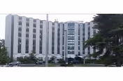Exterior view of a multi-story senior living facility building with multiple windows and a central glass section, surrounded by trees and a parking area in front.