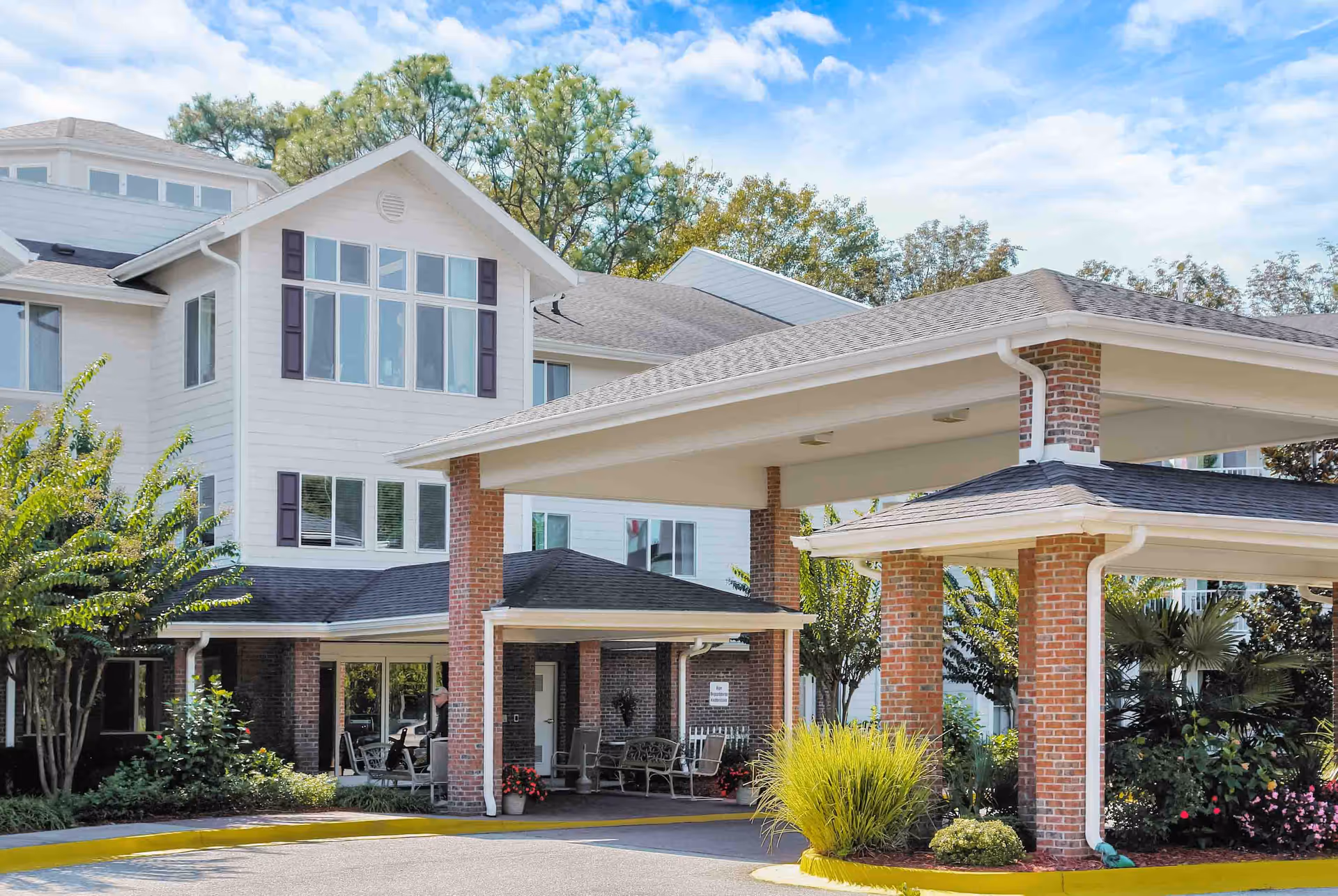 Exterior view of The Woods at Holly Tree Gracious Retirement Living facility showing a covered entrance with brick pillars, surrounded by greenery and trees under a partly cloudy sky.