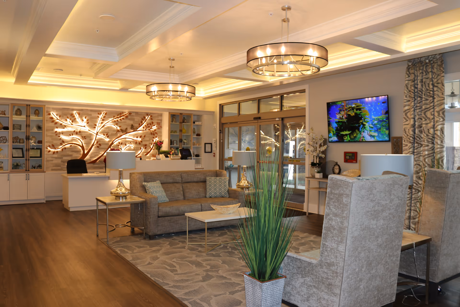 A well-lit senior living facility lobby with a seating area including a sofa, armchairs, and side tables with lamps. The reception desk is in the background with a decorative illuminated tree wall art behind it. There is a large plant in the foreground, a flat-screen TV mounted on the wall, and large glass entrance doors.