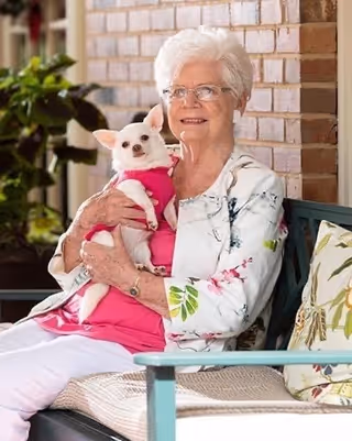An elderly woman with white hair and glasses sitting on a cushioned bench, holding a small white dog dressed in a pink outfit. They are in an outdoor or semi-outdoor setting with a brick wall and some green plants in the background.
