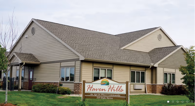 Exterior view of Haven Hills Assisted Living and Memory Care facility, a single-story building with beige siding and a brown shingled roof, surrounded by green grass and some small trees. A sign in front reads 'Haven Hills, An Age in Place Community, 215 Dale Drive.'