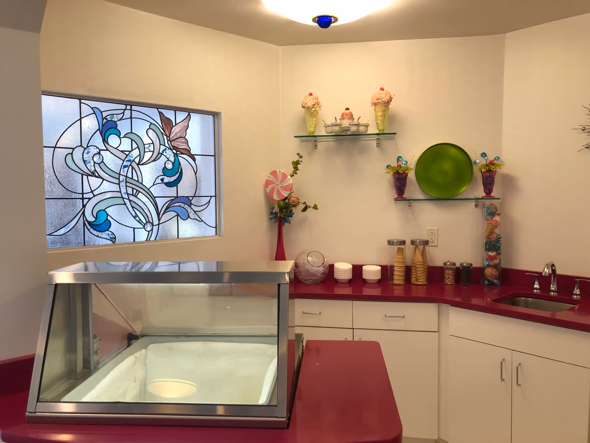 Interior view of a small ice cream or dessert serving area with a red countertop and white cabinets. There is an empty glass display freezer on the counter, a stained glass window with a floral design on the left wall, and decorative items including ice cream cone sculptures, bowls, and containers with cones and toppings on glass shelves and the counter. A small sink with a faucet is on the right side.