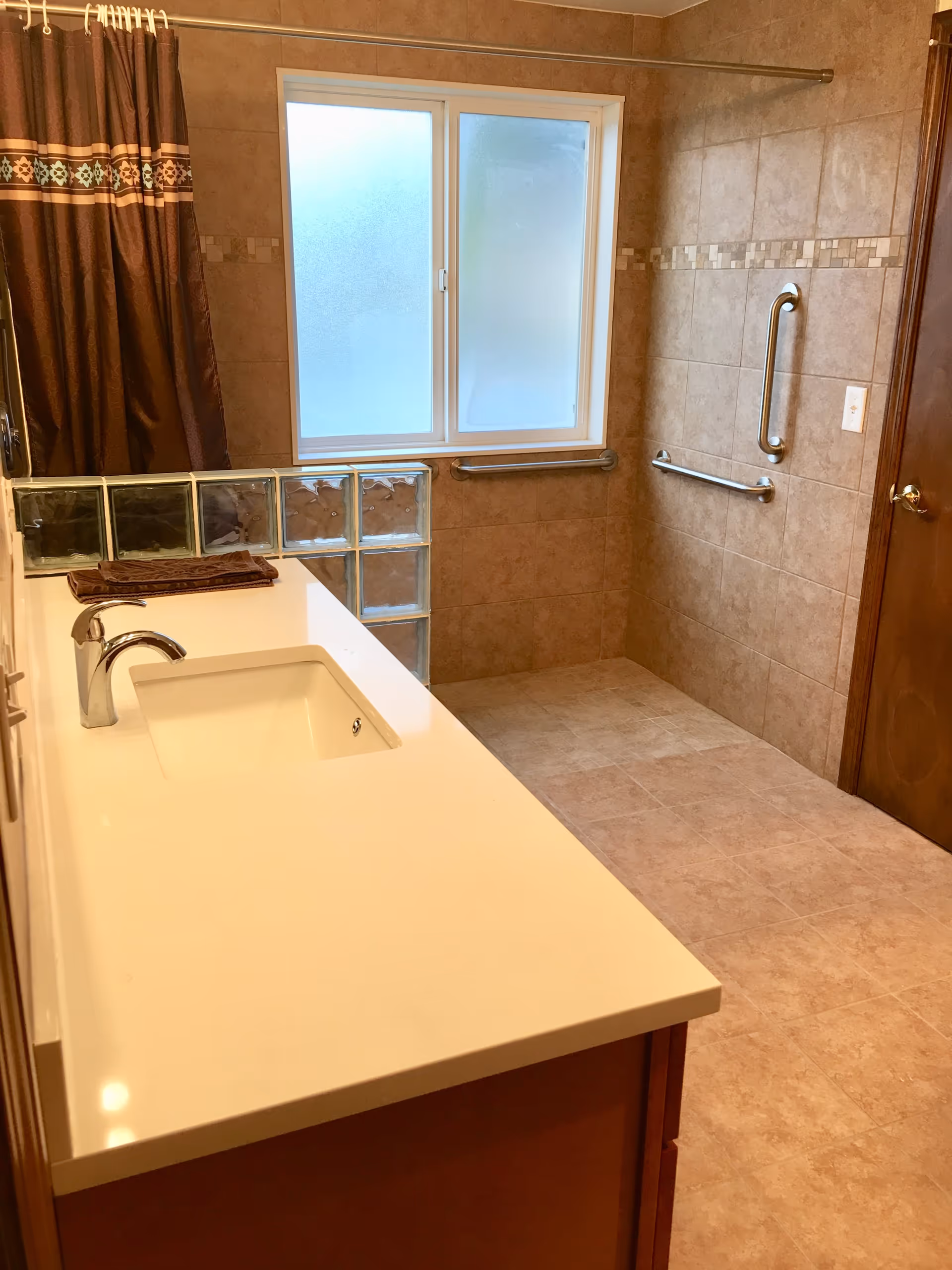 Tiled accessible bathroom with a long vanity sink, grab bars, glass block partition, shower curtain and frosted window.