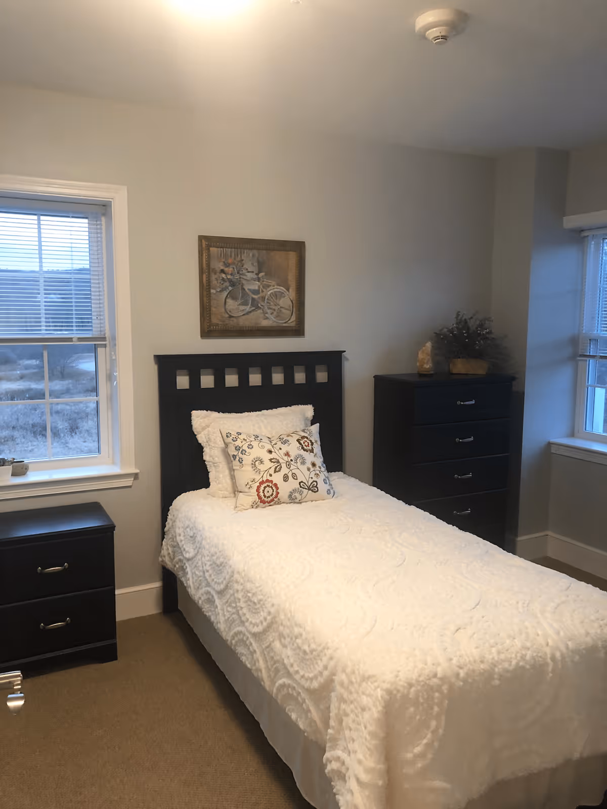 A small bedroom with a single bed covered in a white textured bedspread and two decorative pillows. The bed has a dark wooden headboard. To the left of the bed is a dark wooden nightstand with two drawers, and to the right is a matching dark wooden chest of drawers with decorative items on top. There are two windows with blinds, one on the left wall and one on the right wall. A framed picture of a bicycle with flowers hangs on the wall above the bed.