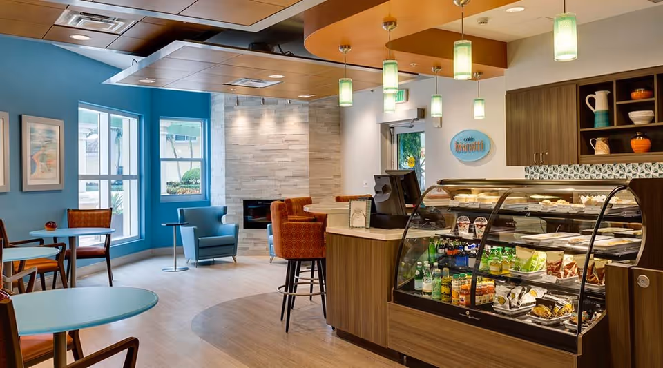 A cozy cafe area inside a senior living facility with a display case filled with beverages and snacks, a counter with a cash register, several tables and chairs, blue walls with framed artwork, and a seating area with armchairs near a modern fireplace.