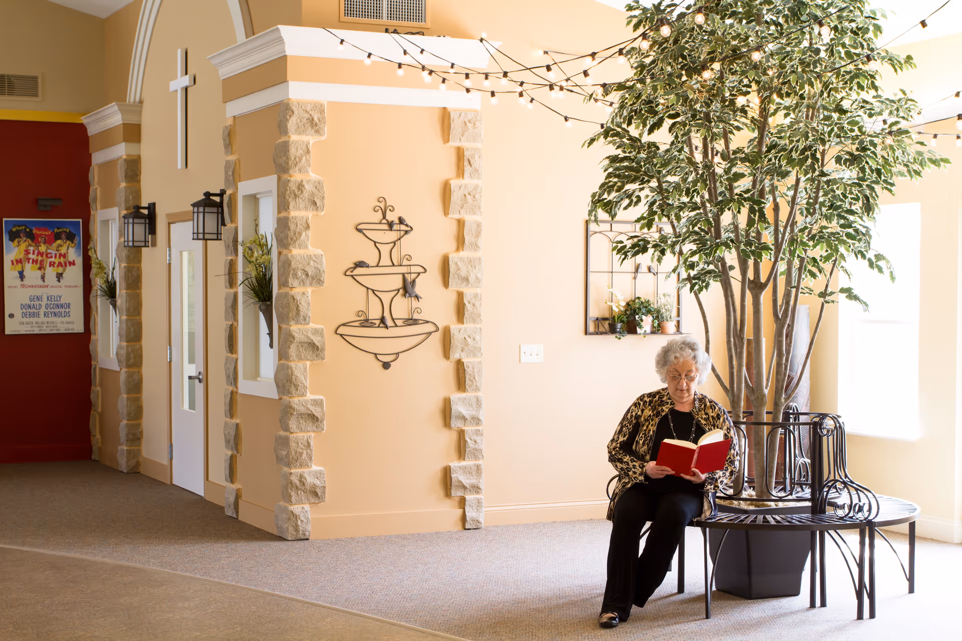 An older woman sits on a circular bench around a potted tree in a bright common room reading a book near decorative wall features.