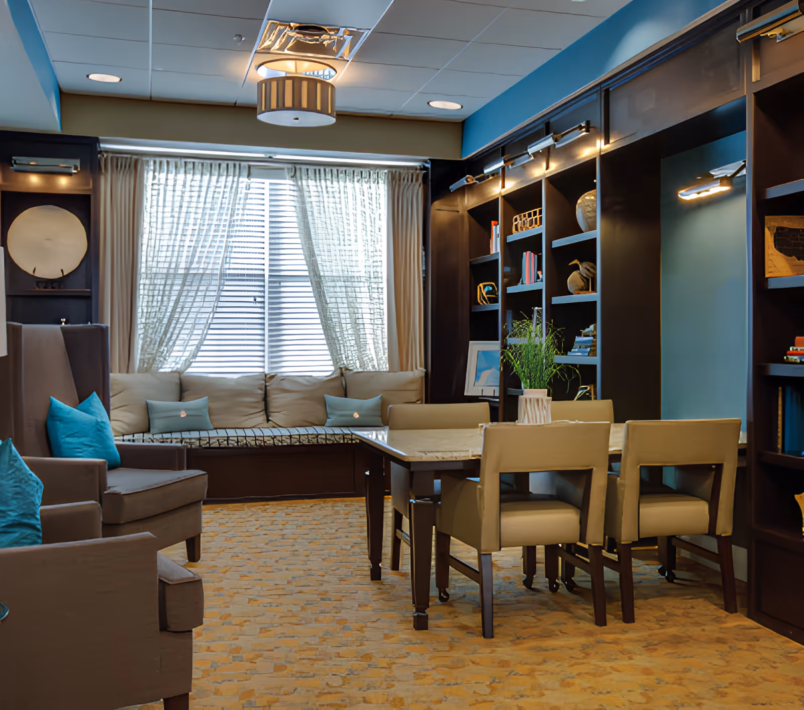 Sunlit communal lounge with a table and chairs, armchairs, built-in bookshelves and a window seat.