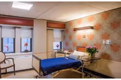 A hospital-style bedroom with a single bed covered in a blue blanket, a bedside table with a vase of flowers, two chairs near the windows, and floral patterned wallpaper on one wall.