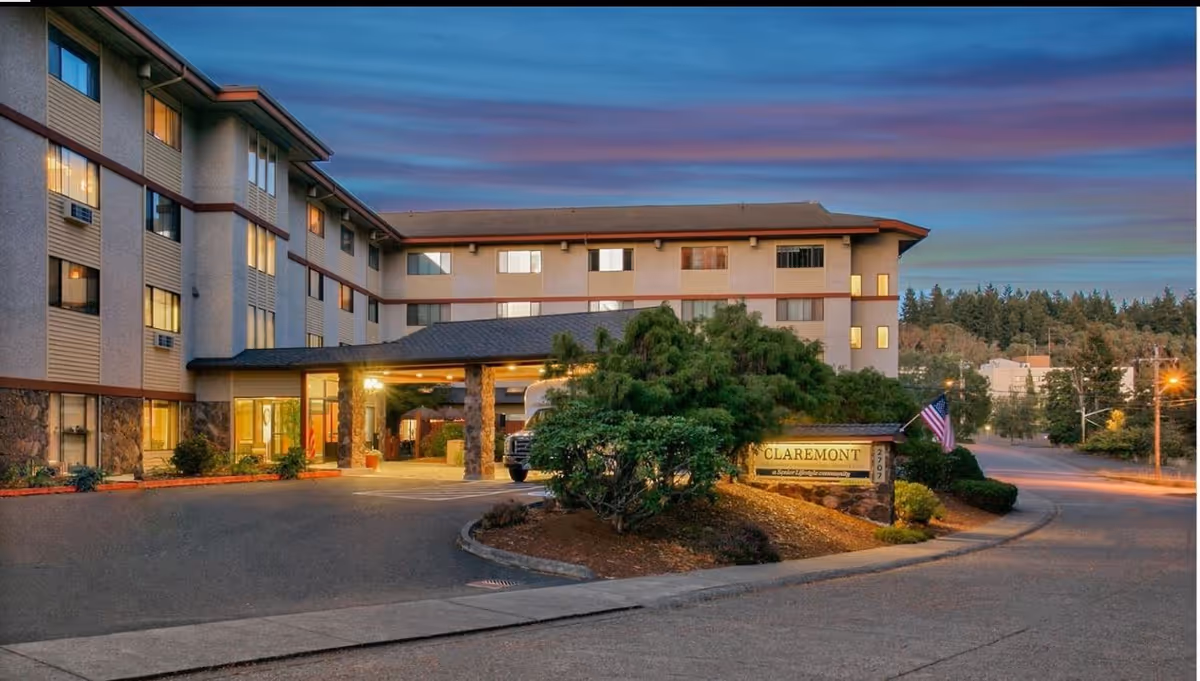 Front entrance of a multi-story senior living building at dusk with a lit porte-cochere, landscaping, and a sign reading 'Claremont'.