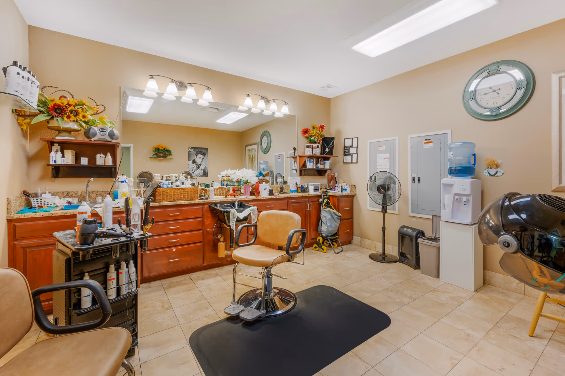 Interior view of a hair salon area in a senior living facility with a salon chair on a black mat, a hair washing sink, wooden cabinets with various hair care products, a large mirror with lights above it, a standing fan, a water dispenser, and a wall clock.