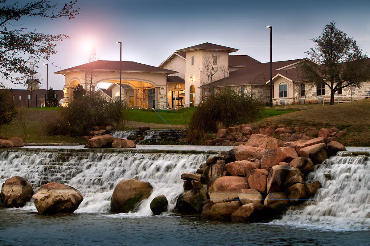 Exterior view of Volante Senior Living of Round Rock at dusk, showing a large building with stone and stucco walls, a covered entrance, surrounding trees, grassy areas, and a cascading waterfall over rocks in the foreground.