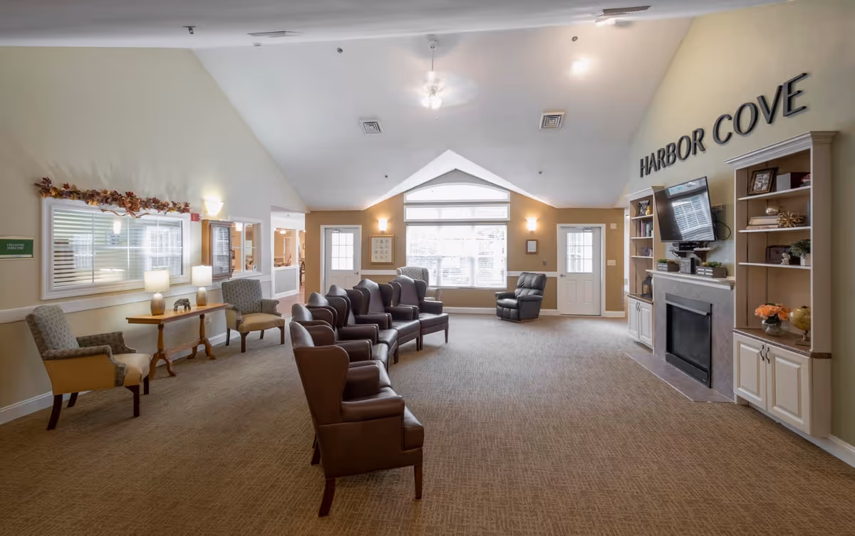 A spacious and well-lit common area in Harbor Cove Memory Care featuring multiple brown leather chairs arranged in rows facing a fireplace with a mounted TV above it. The room has beige walls, carpeted floor, and large windows letting in natural light. There are built-in shelves on either side of the fireplace with decorative items, and additional seating with upholstered chairs and small tables along the left wall. The ceiling is vaulted with a ceiling fan in the center.