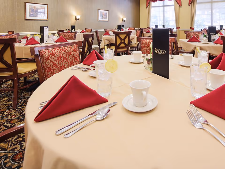 Dining room with round tables covered in beige tablecloths, set with red folded napkins, white cups and saucers, glasses of water with lemon slices, silverware, and black menus labeled 'Legend Senior Living'. The room has patterned carpet, upholstered chairs, framed artwork on the walls, and large windows with red curtains allowing natural light.