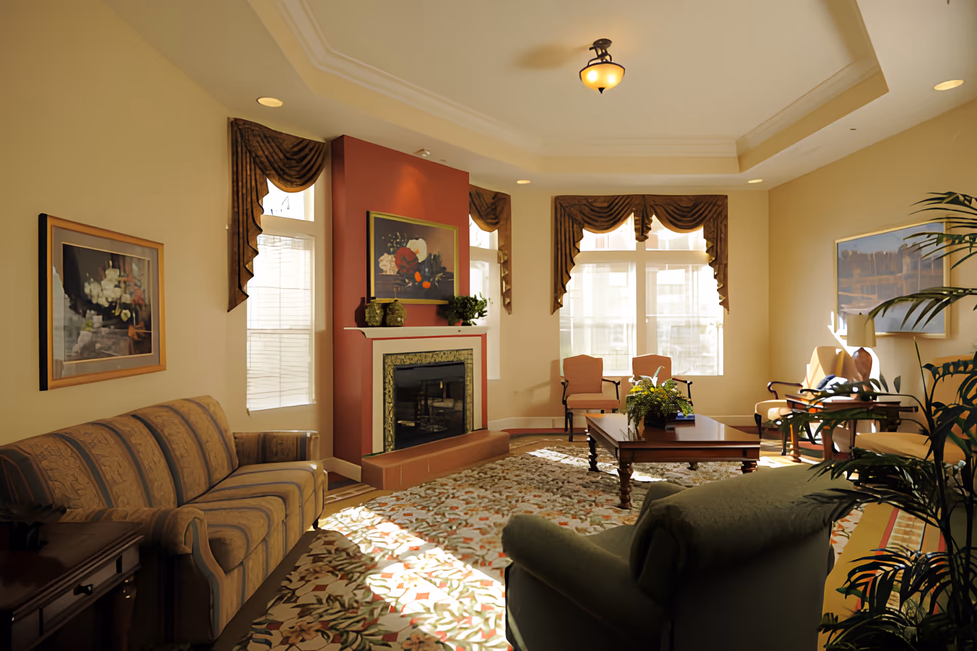 A cozy senior living room with a patterned sofa, armchairs, a wooden coffee table with a plant, a fireplace with decorative vases and a painting above it, large windows with brown drapes, and a floral area rug covering the floor.