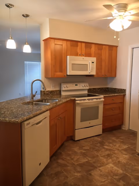 Modern kitchen with wooden cabinets, granite countertops, a white dishwasher, a white stove with oven, a white microwave above the stove, a double sink with a curved faucet, two hanging pendant lights, and a ceiling fan with lights.