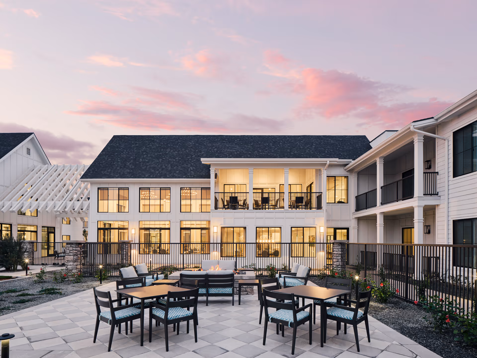 Outdoor courtyard with patio tables, chairs and a fire pit in front of a two-story senior living building at sunset.
