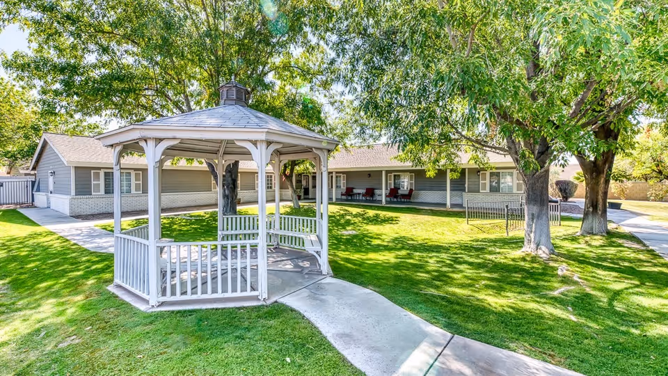 A well-maintained outdoor garden area at the Cottages at Peoria featuring a white wooden gazebo with benches inside, surrounded by green grass, trees providing shade, and a paved walkway leading to the gazebo and the building in the background.