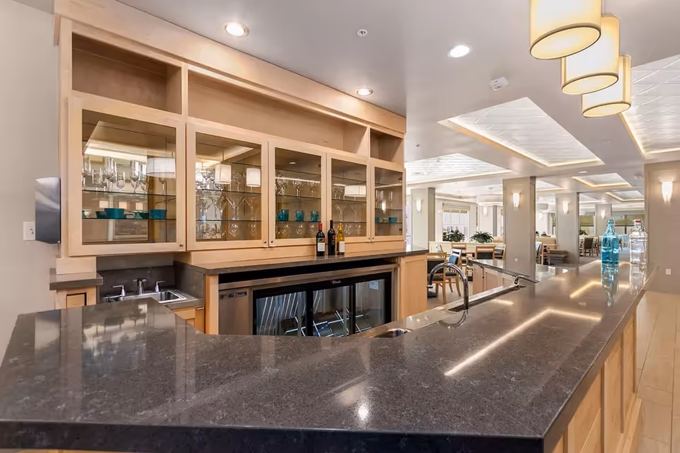 Interior view of a modern kitchen and dining area in a senior living facility. The kitchen features a large dark granite countertop with a built-in sink and a glass-front cabinet displaying glassware and blue bowls. There are three pendant lights hanging above the counter. In the background, there are multiple dining tables and chairs arranged in a spacious, well-lit room with ceiling lights and wall sconces.