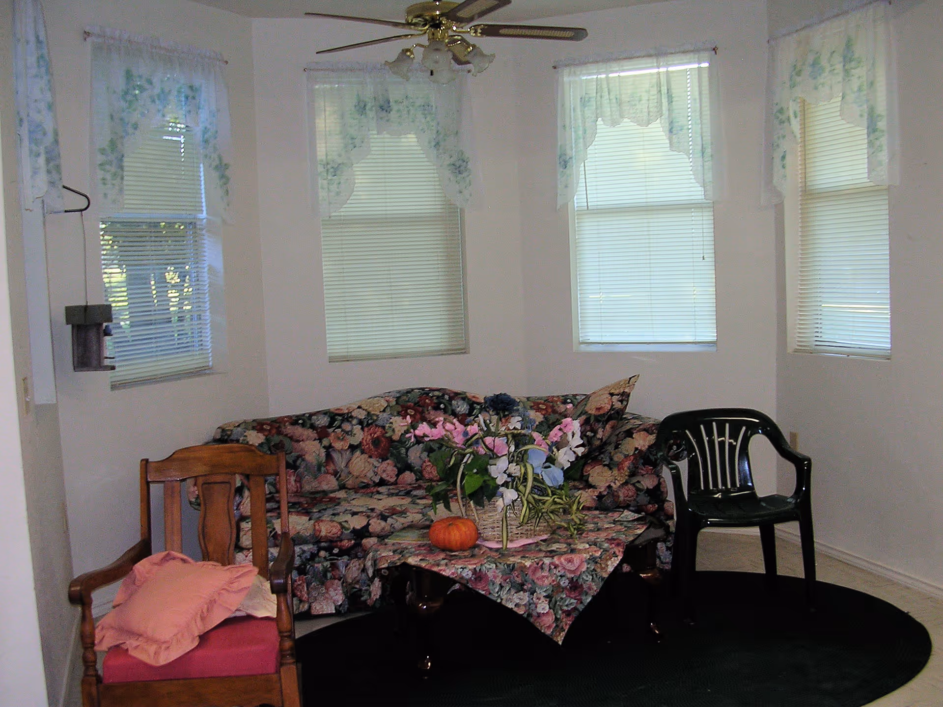 A small living room with a floral sofa, wooden chair, black plastic chair, and a table with flowers near several windows.