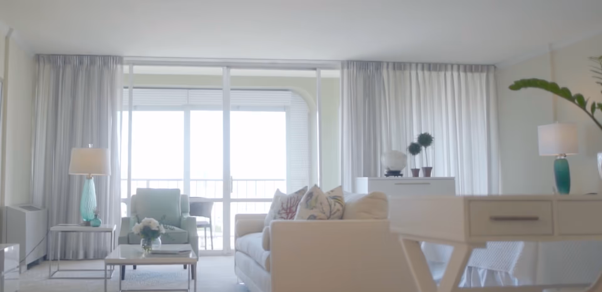A bright and airy living room with a white sofa adorned with patterned pillows, a light green armchair, a glass coffee table with a flower vase, two turquoise lamps on side tables, white curtains covering large sliding glass doors leading to a balcony, and a white desk with drawers in the foreground.