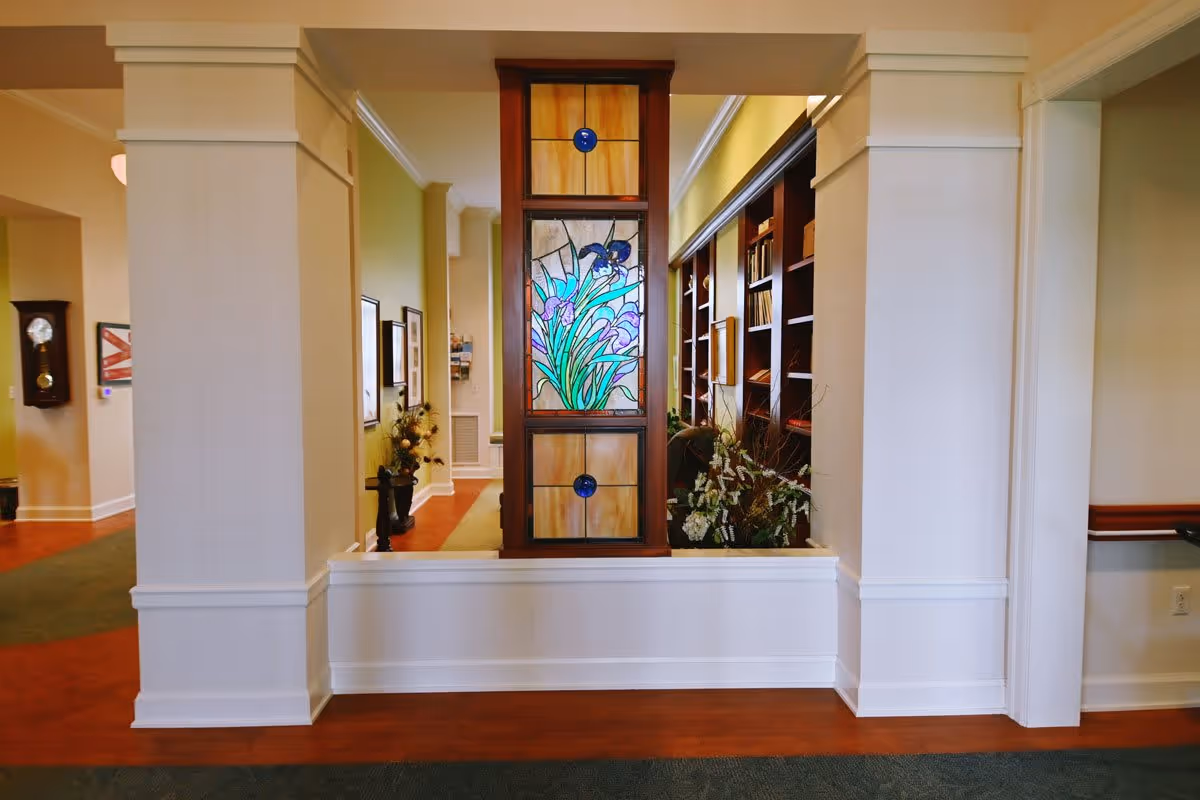 Interior view of a senior living facility hallway with a decorative stained glass panel featuring purple flowers in the center. The hallway has light-colored walls, wooden flooring, and built-in bookshelves on the right side filled with books and decorative items. There is a grandfather clock and framed pictures on the left wall.