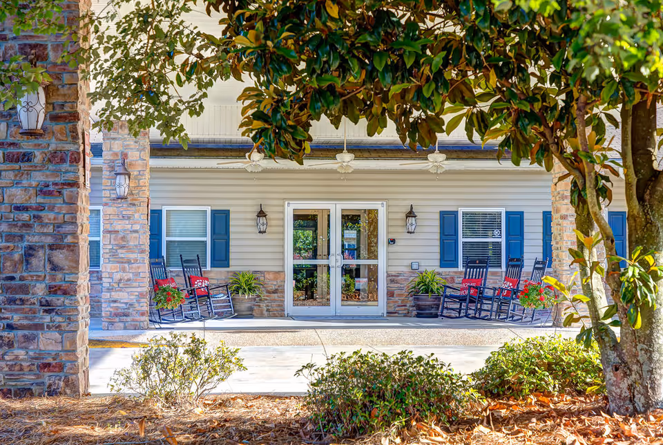 Front entrance of a senior living facility with a covered porch, stone columns, double glass doors, rocking chairs and potted plants.