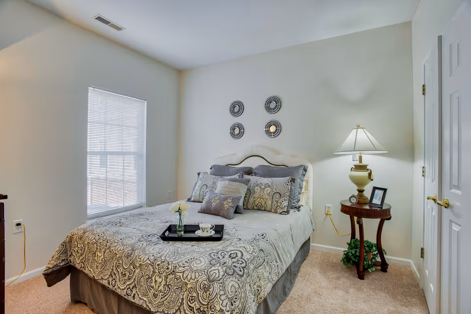 A cozy bedroom with a neatly made bed featuring patterned bedding and multiple pillows. There is a small round wooden side table with a lamp, a framed photo, and a small clock. Four decorative circular mirrors are mounted on the wall above the bed. A window with blinds allows natural light into the room, and a closed white door is visible on the right side.