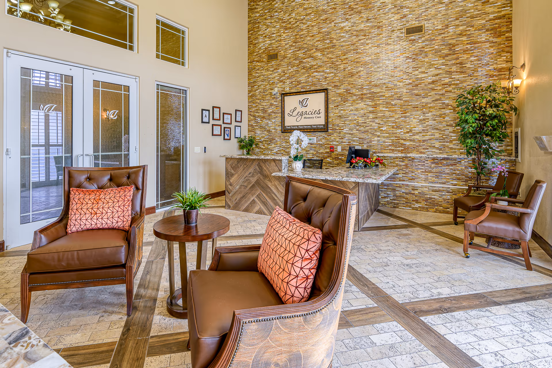 A senior living facility reception area with a tiled accent wall behind a granite countertop desk. Two brown leather chairs with orange patterned pillows and a small round wooden table with a plant are in the foreground. Additional seating and plants are arranged around the room, with glass double doors and framed certificates on the wall.