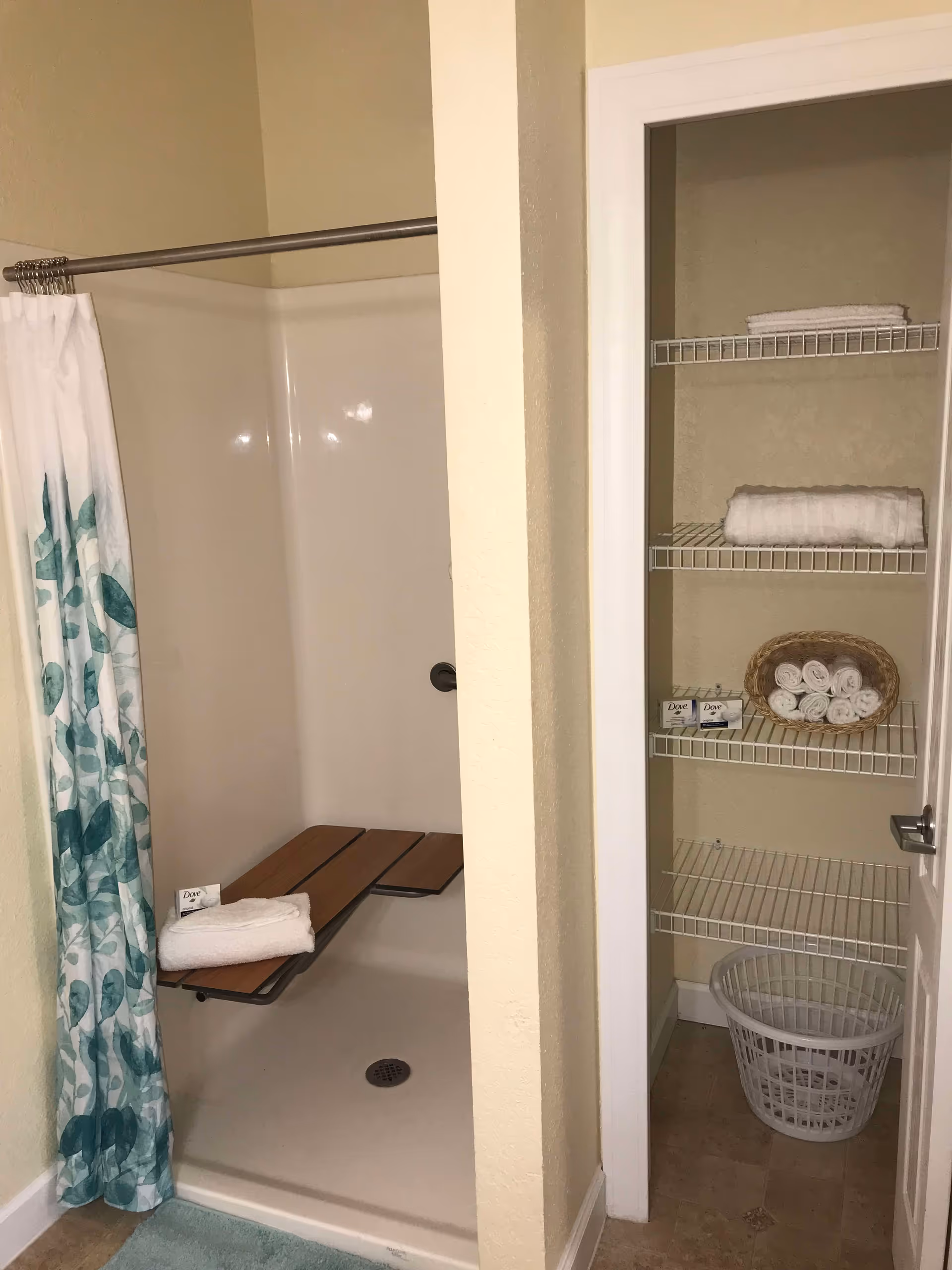 Shower with a folding wooden bench and patterned curtain beside an open linen closet holding rolled towels and a laundry basket.