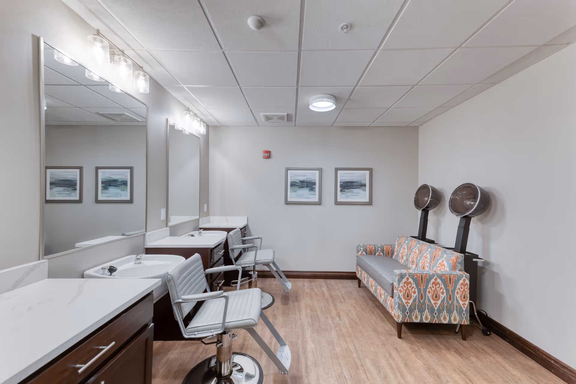 Interior view of a salon area in a senior living facility featuring two styling chairs in front of large mirrors and sinks, a patterned sofa with two vintage hair dryers mounted on stands, light wood flooring, and two framed abstract paintings on the wall.