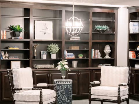 A cozy interior room featuring two cushioned armchairs with wooden frames facing each other, a small round table with a floral arrangement between them, and a large dark wood built-in bookshelf in the background filled with decorative items, books, and plants. A modern chandelier hangs from the ceiling above the table.