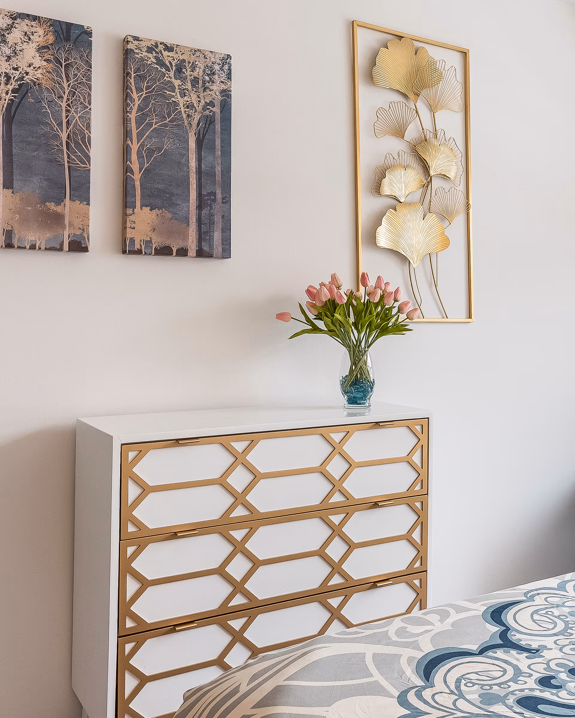 A white dresser with gold geometric trim sits against a white wall. On top of the dresser is a glass vase with pink tulips. Above the dresser, there are two pieces of wall art: one is a pair of paintings depicting trees with gold accents, and the other is a gold metal wall sculpture of stylized leaves. Part of a bed with a blue and gray patterned bedspread is visible in the foreground.