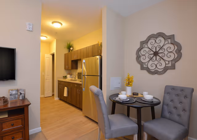 Interior view of a senior living facility apartment at Anthem Lakes showing a small dining area with a round table set for two, two gray upholstered chairs, and a decorative wall piece. In the background, there is a kitchenette with wooden cabinets, a stainless steel refrigerator, and a countertop with a few items. A hallway with a closed door is visible beyond the kitchenette.
