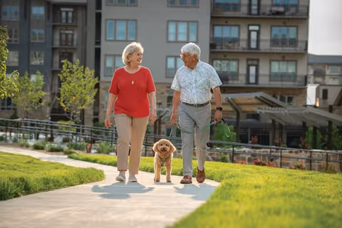 An elderly couple walking a small dog on a paved path in a grassy outdoor area with a multi-story residential building in the background.
