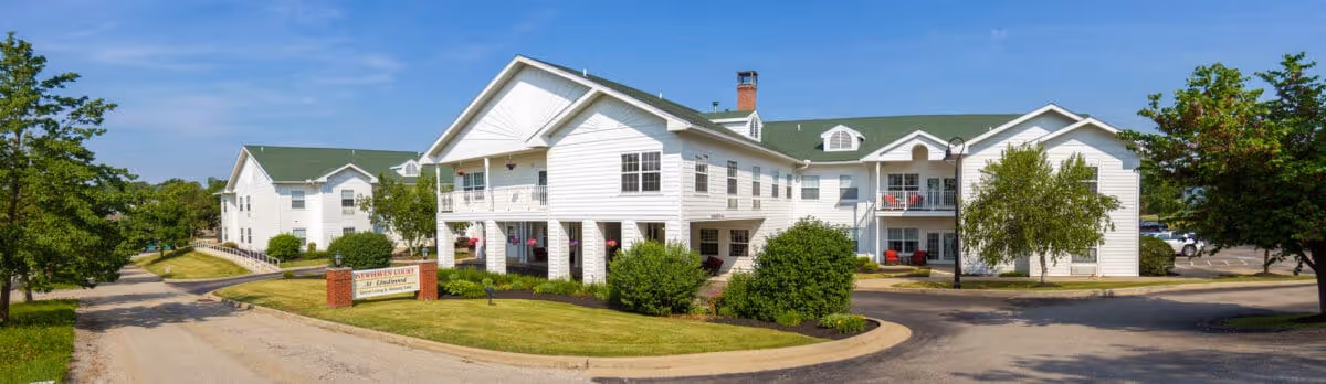 White two-story senior living building with green roofs, balconies, a circular driveway, and landscaped lawns.