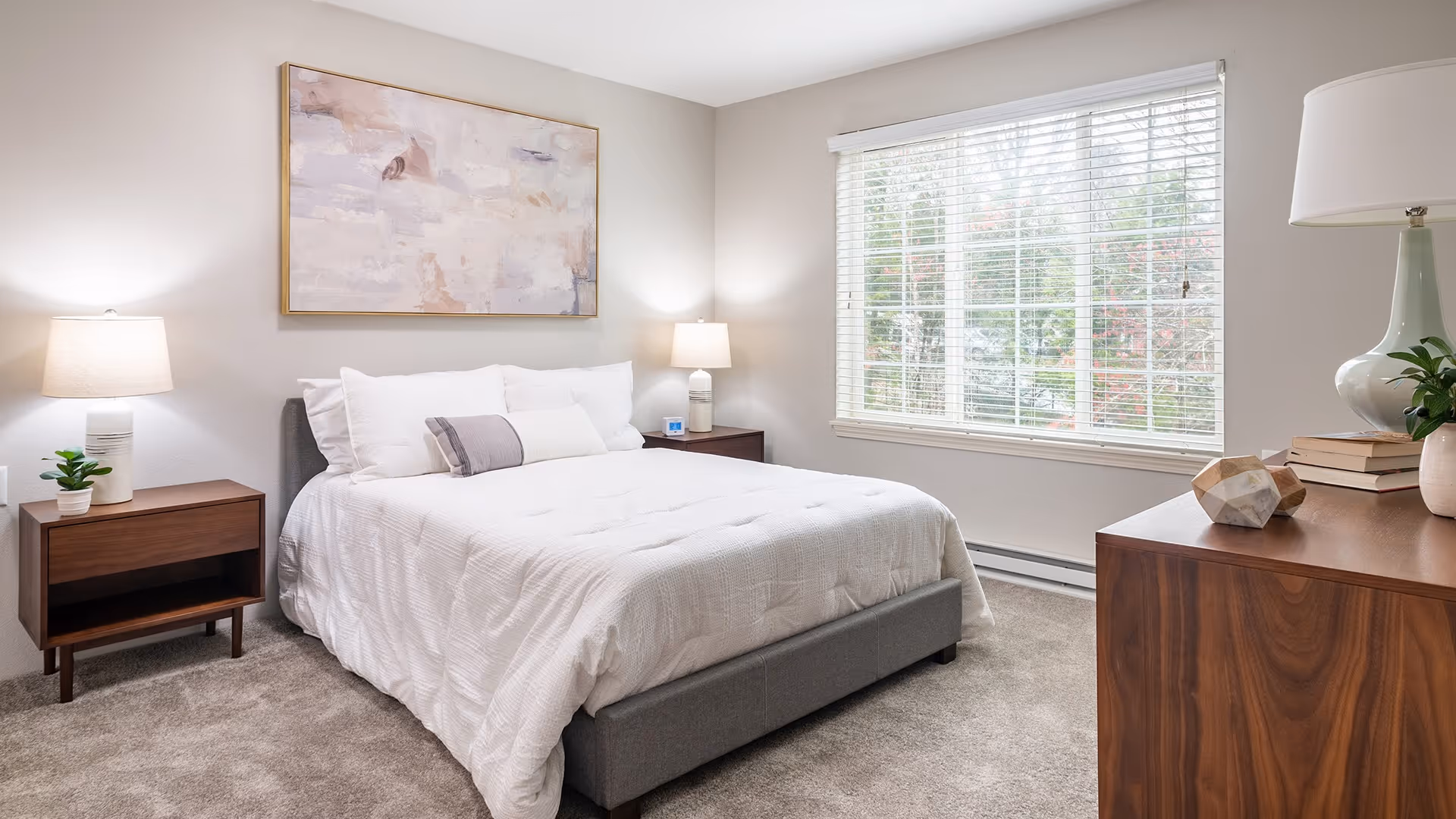 A bright and clean bedroom with a large window covered by white blinds. The room features a neatly made bed with white bedding and multiple pillows, flanked by two wooden nightstands each with a lamp and small decorative items. A wooden dresser with a lamp, books, and a plant is positioned near the window. A large abstract painting hangs above the bed on the light-colored wall.