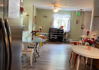 Interior view of a dining area in an assisted living facility with several round tables and chairs. The room has light green walls, a window with white curtains, a ceiling fan, and a black chest of drawers near the window. There are flower arrangements on the tables and a refrigerator visible on the left side.