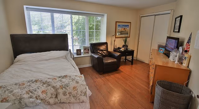 A bedroom with a large window covered by white blinds, a bed with floral bedding, a black leather armchair, a wooden nightstand with a lamp and framed photos, a wooden dresser with a TV on top, and a laundry basket on the floor.