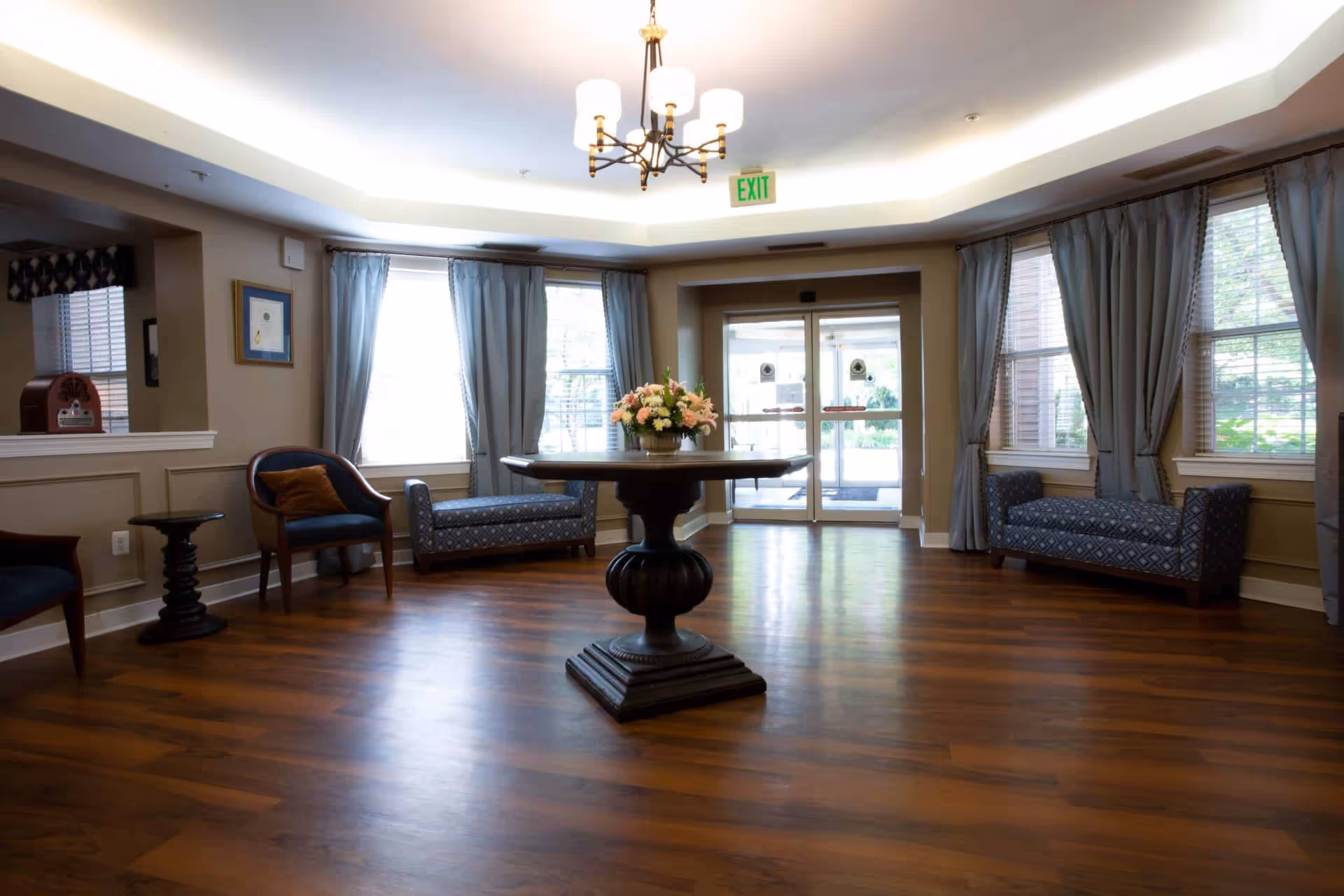 A spacious and well-lit common area with wooden flooring, a round wooden table with a flower arrangement in the center, two patterned blue benches near windows with light blue curtains, and two blue chairs with brown cushions. There is a chandelier hanging from the ceiling and glass double doors leading outside.