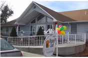 Exterior view of a single-story assisted living facility with a sloped roof, a white railing around a raised porch area, and colorful balloons attached near the entrance. A sign in front reads 'Aspen Leaf Assisted Living'.