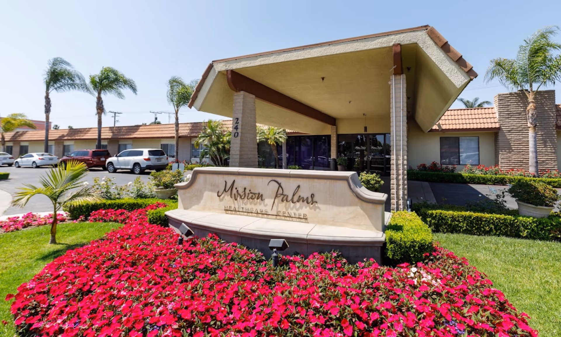 Entrance of Mission Palms Healthcare Center with a covered driveway, a large sign displaying the facility name, surrounded by vibrant red flowers and palm trees under a clear blue sky.