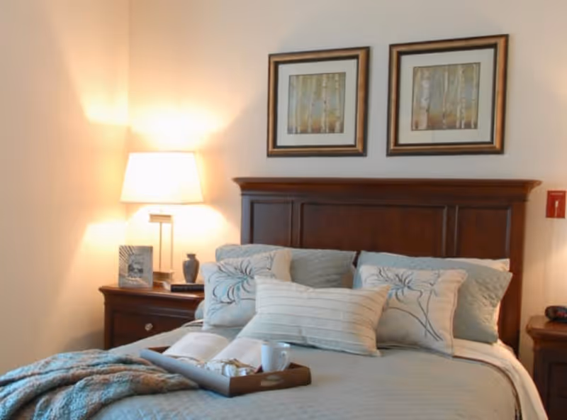 A cozy bedroom with a wooden headboard, a bed neatly made with multiple pillows and a light blue quilt. There is a wooden nightstand on the left side with a table lamp, a small vase, and a framed photo. Two framed pictures hang on the wall above the headboard.
