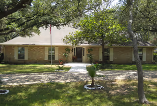 Single-story brick building with a centered entrance, flagpole, shaded front yard, and a paved walkway leading to the door.