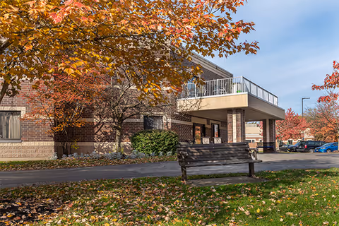 Exterior view of Elderwood Assisted Living at Cheektowaga showing a brick building with a covered entrance, a bench on a grassy area with fallen autumn leaves, and trees with orange and red foliage under a clear blue sky.