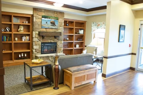 A cozy living room area in The Gables of Canton featuring a stone fireplace with a mounted picture above it, wooden built-in bookshelves on either side, a gray sofa, a wooden bench, a side table with a lamp, and hardwood flooring.