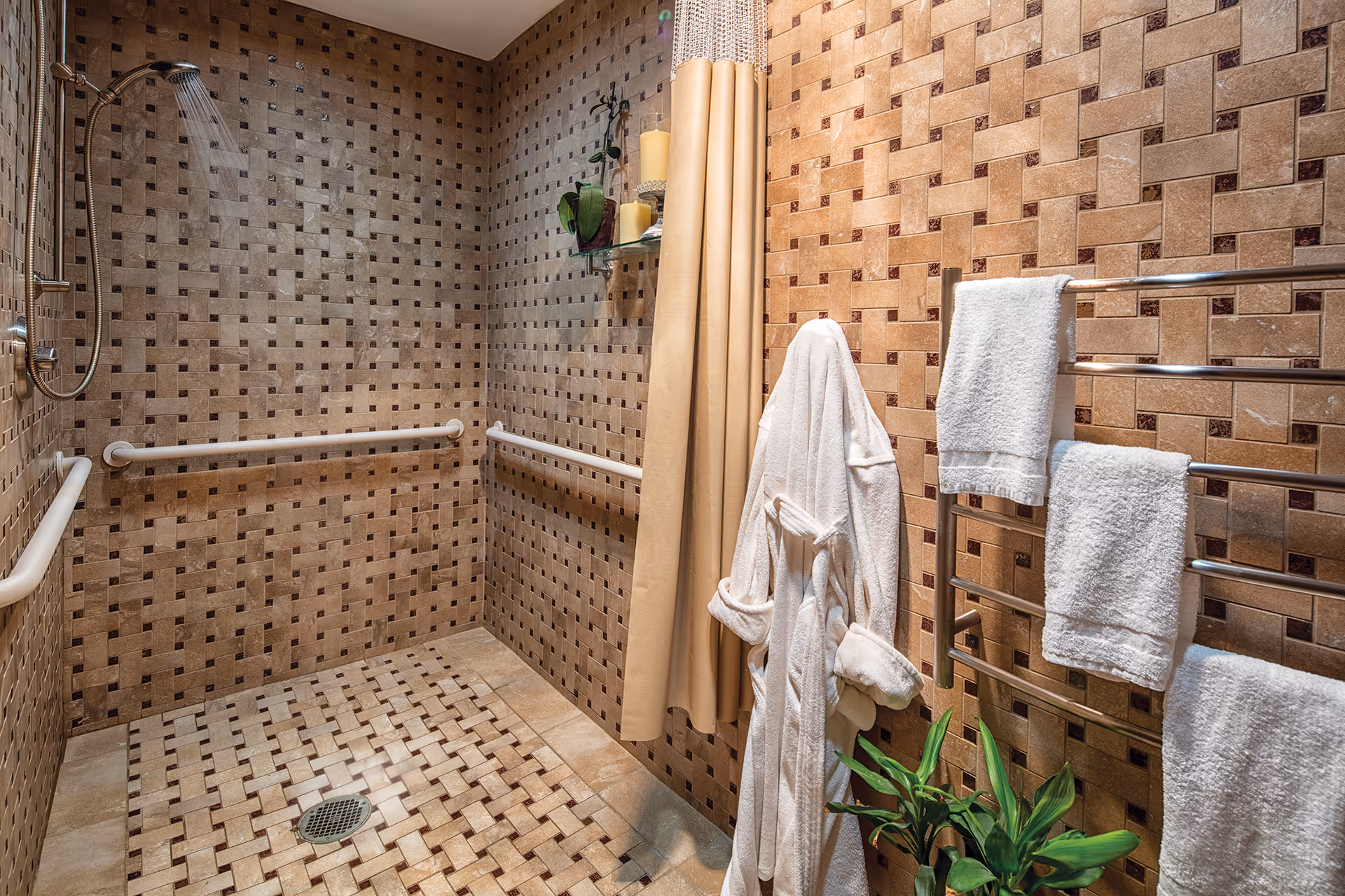 Walk-in tiled bathroom shower with grab bars, a wall-mounted showerhead, hanging bathrobe, towel rack, and a small plant.