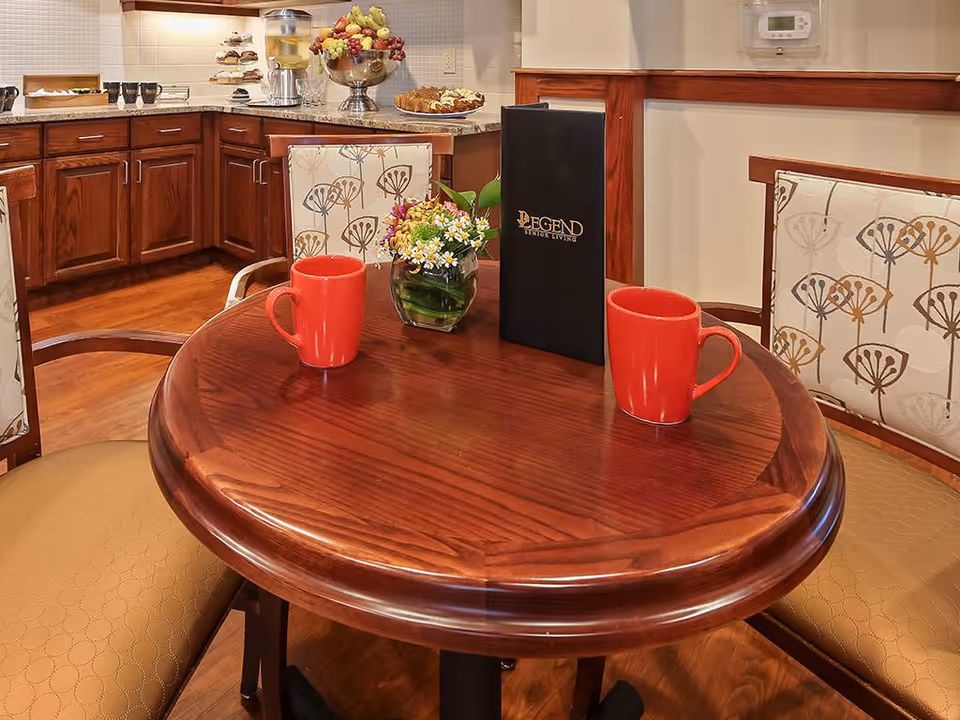 A round wooden table with two red mugs, a small vase of flowers, and a black menu with the text 'Legend Senior Living' on it. The table is surrounded by cushioned chairs with patterned upholstery. In the background, there is a kitchen counter with wooden cabinets, a fruit bowl, a beverage dispenser, and plates of pastries.