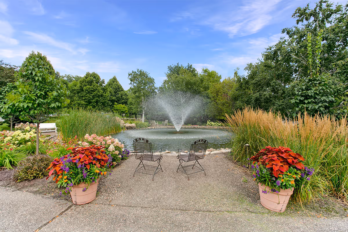 A peaceful outdoor garden area featuring a small pond with a central water fountain spraying water upwards. Two metal chairs face the pond, surrounded by lush greenery, colorful flowers in large pots, and tall grasses under a partly cloudy blue sky.